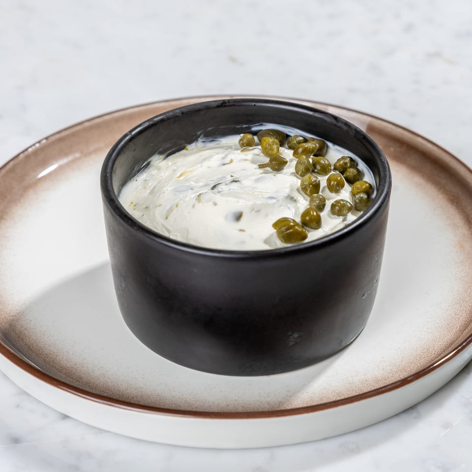 Creamy white dip with green capers in a black bowl on a two-toned plate.
