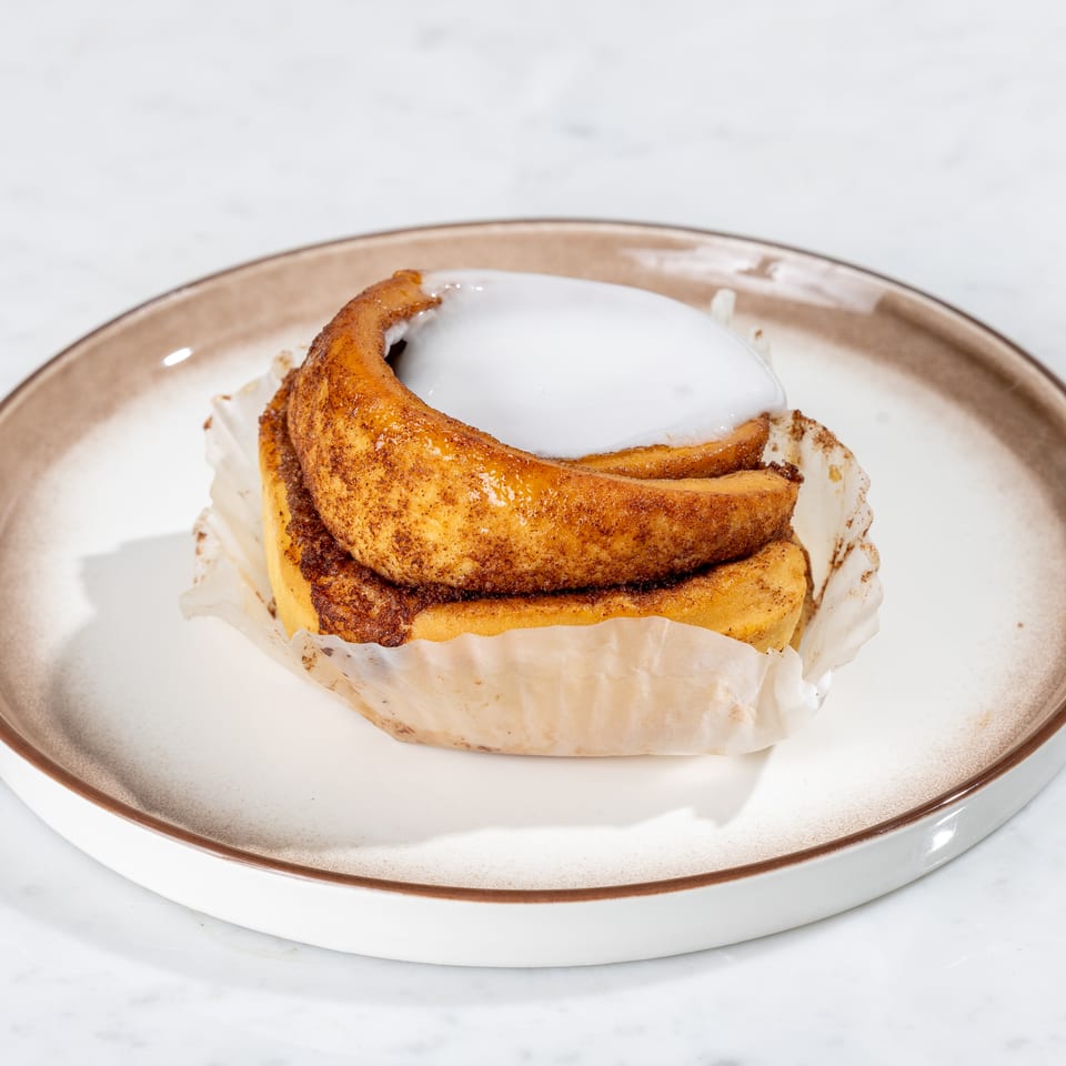 A single cinnamon roll with white frosting, wrapped in paper, on a white and brown plate.